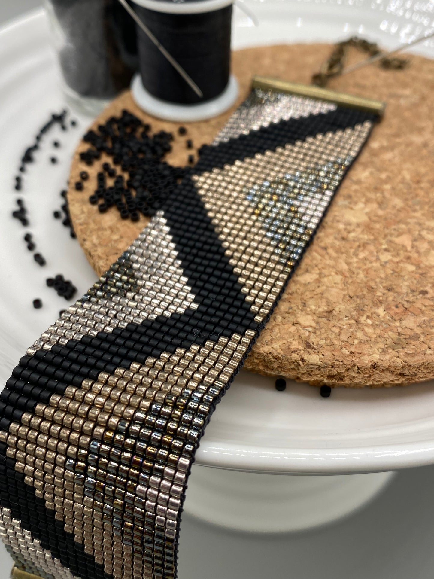 Close up of a black and gold beaded bracelet with a chevron pattern on a white and cork plate with a blurred background.
