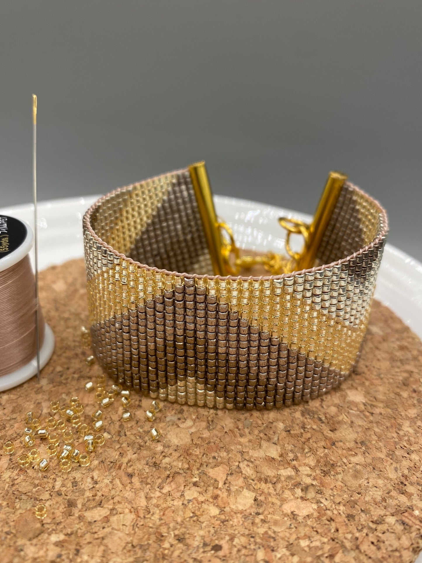 Gold beaded bracelet with a chevron pattern on a white and cork plate with a blurred background.