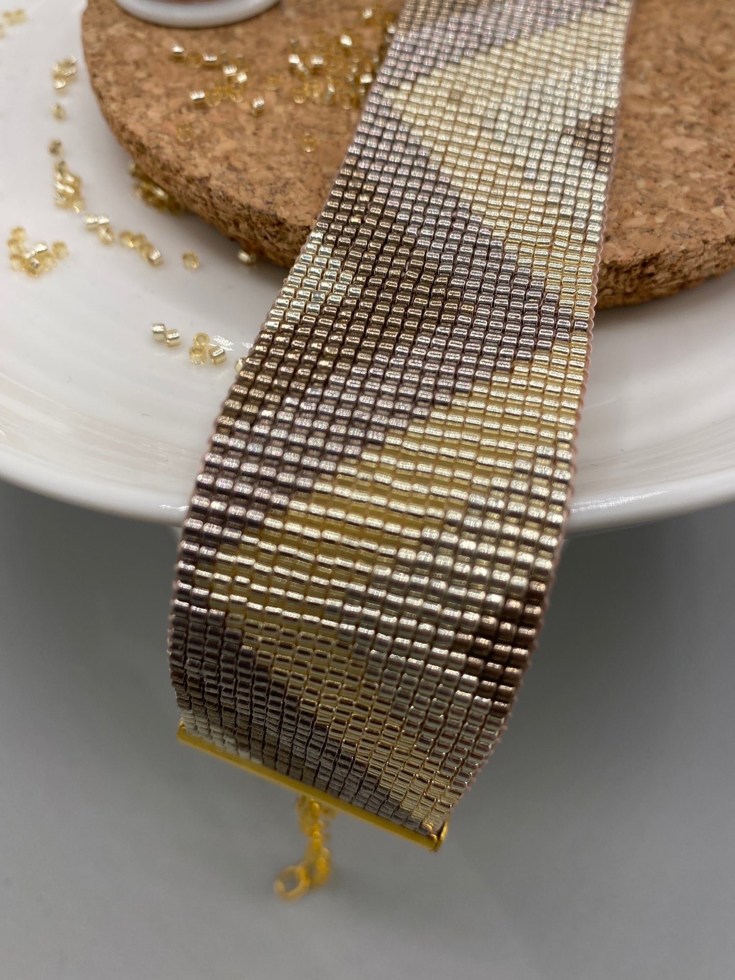 Close up of a gold beaded bracelet with a chevron pattern on a white and cork plate with a blurred background.