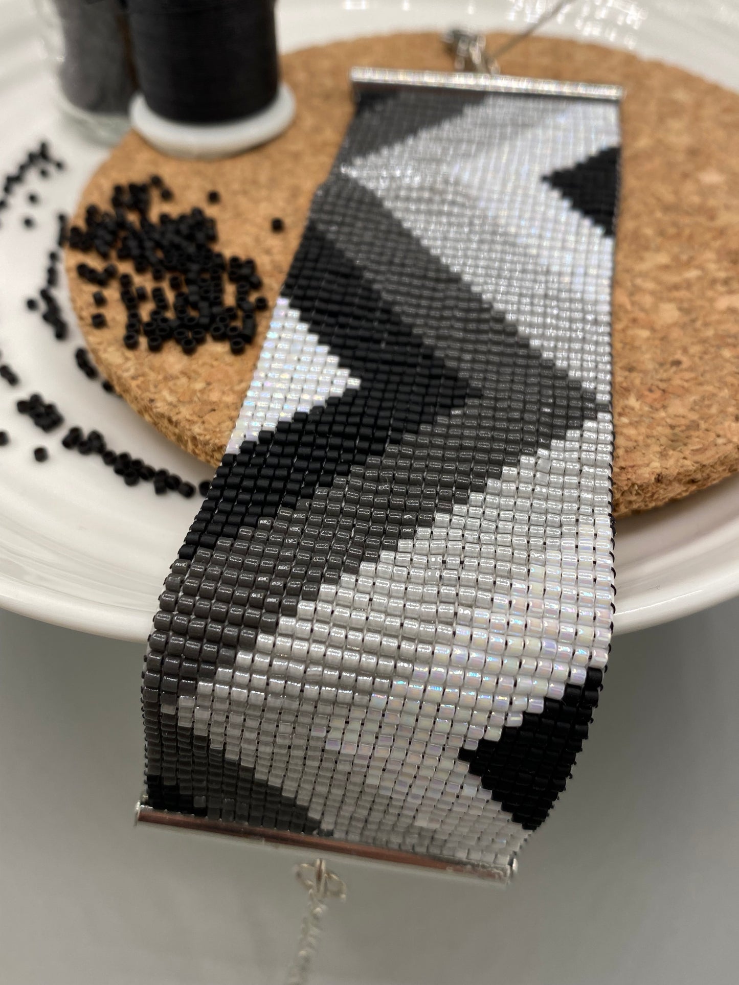Close-up of a black, grey, and white beaded bracelet with chevron pattern on a cork and white plate.