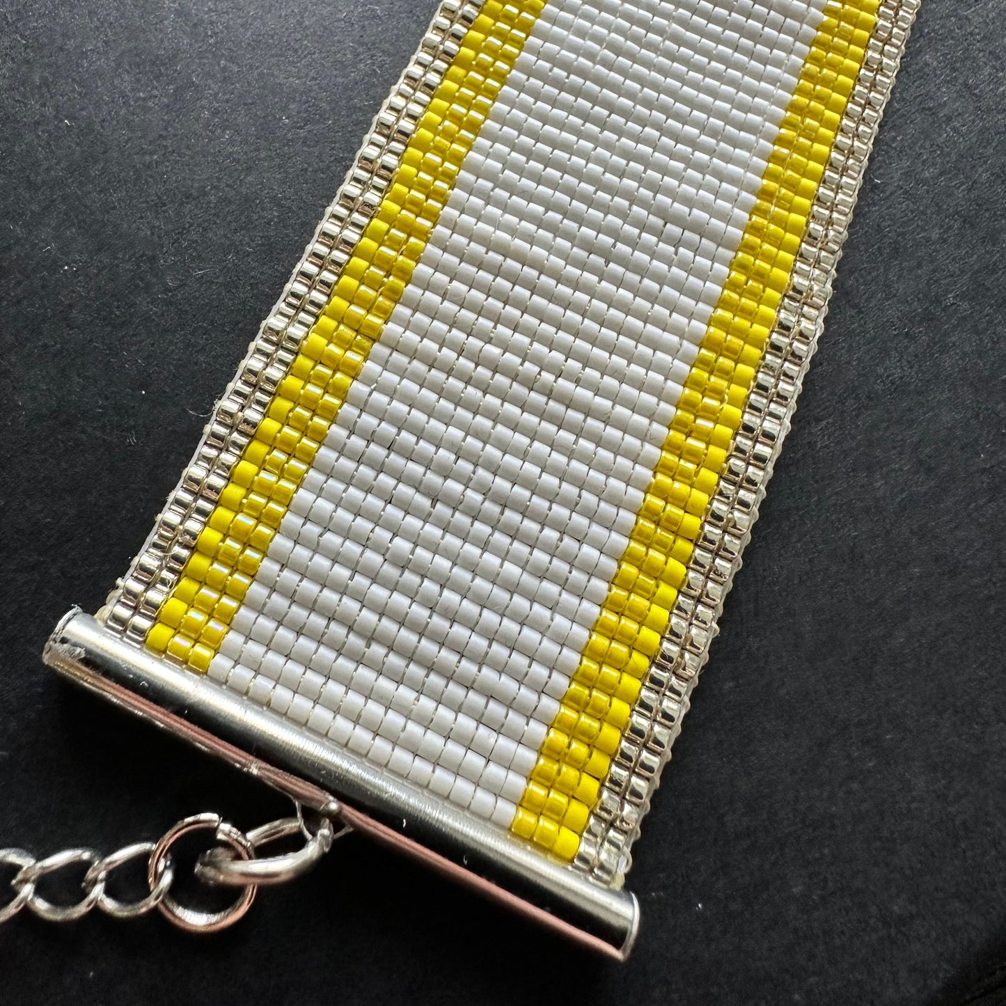 Close up of a beaded bracelet with yellow and gold stripes on edge, white in the middle on a dark surface.