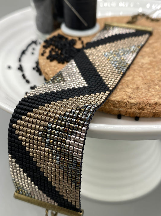 Black and gold beaded bracelet with a chevron pattern on a white and cork plate with a blurred background.