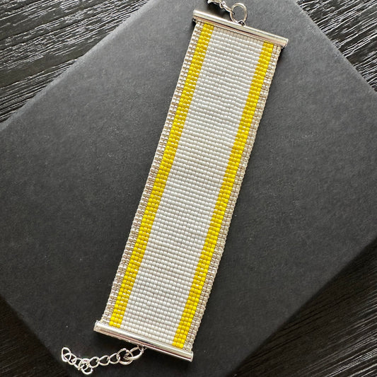 Beaded bracelet with yellow and gold stripes on edge, white in the middle on a dark surface.