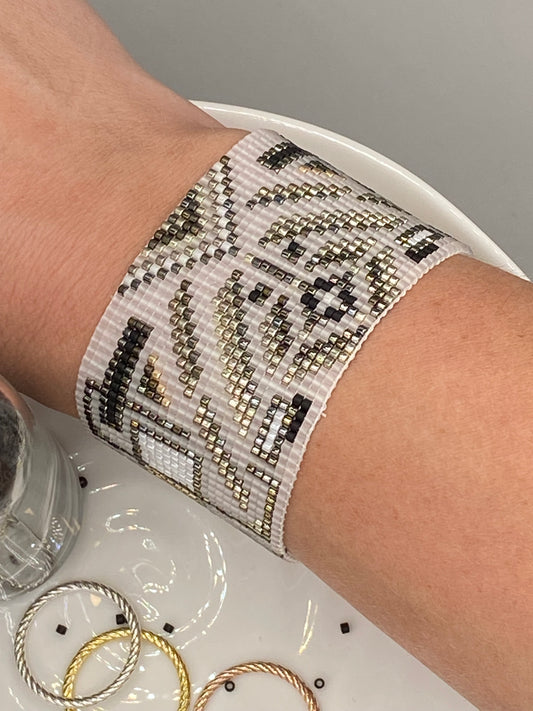 Close-up of a wrist wearing a white and gold bracelet cuff with a pattern, resting on a white plate containing more jewelry in the background.
