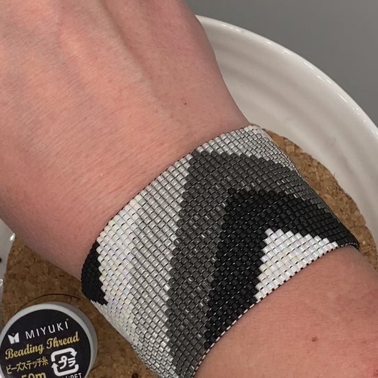 Video of a black, grey, and white beaded bracelet with chevron pattern on a cork and white plate.